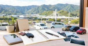 徳島県西部の山並みを望む机に、家族の車管理用ノート、鍵、スマートフォン、予定表を置いた地域カーライフ計画のイメージ